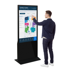 A man in a navy jacket and jeans stands beside a large black freestanding touchscreen kiosk, touching the screen that displays a building directory map. The background is solid white.