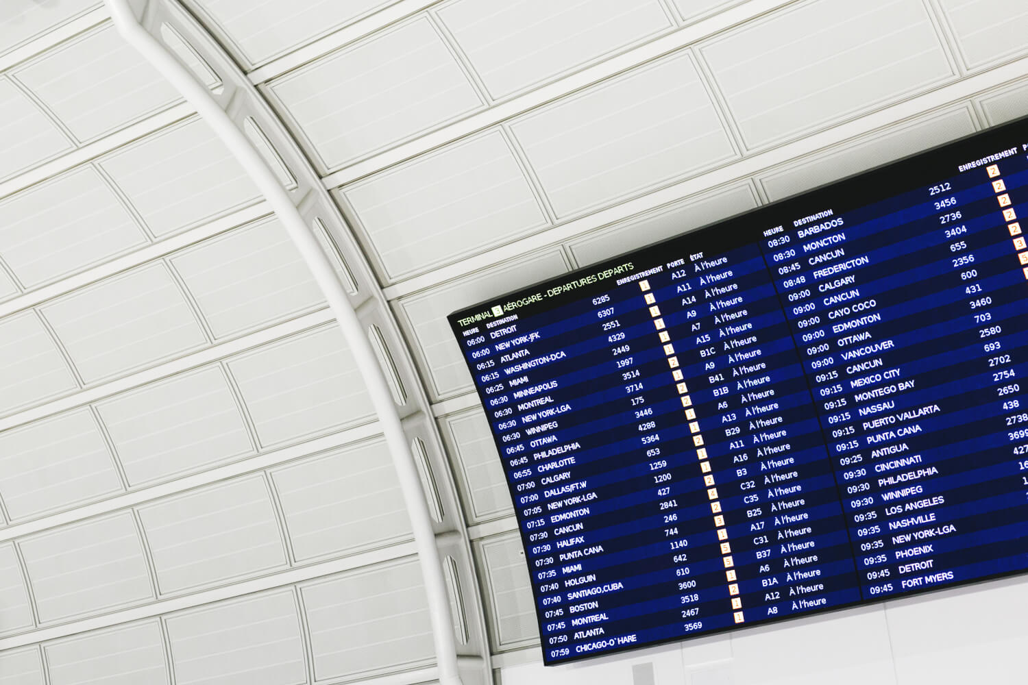 A large airport departure board with blue screens and white text, mounted on a white arched ceiling structure.